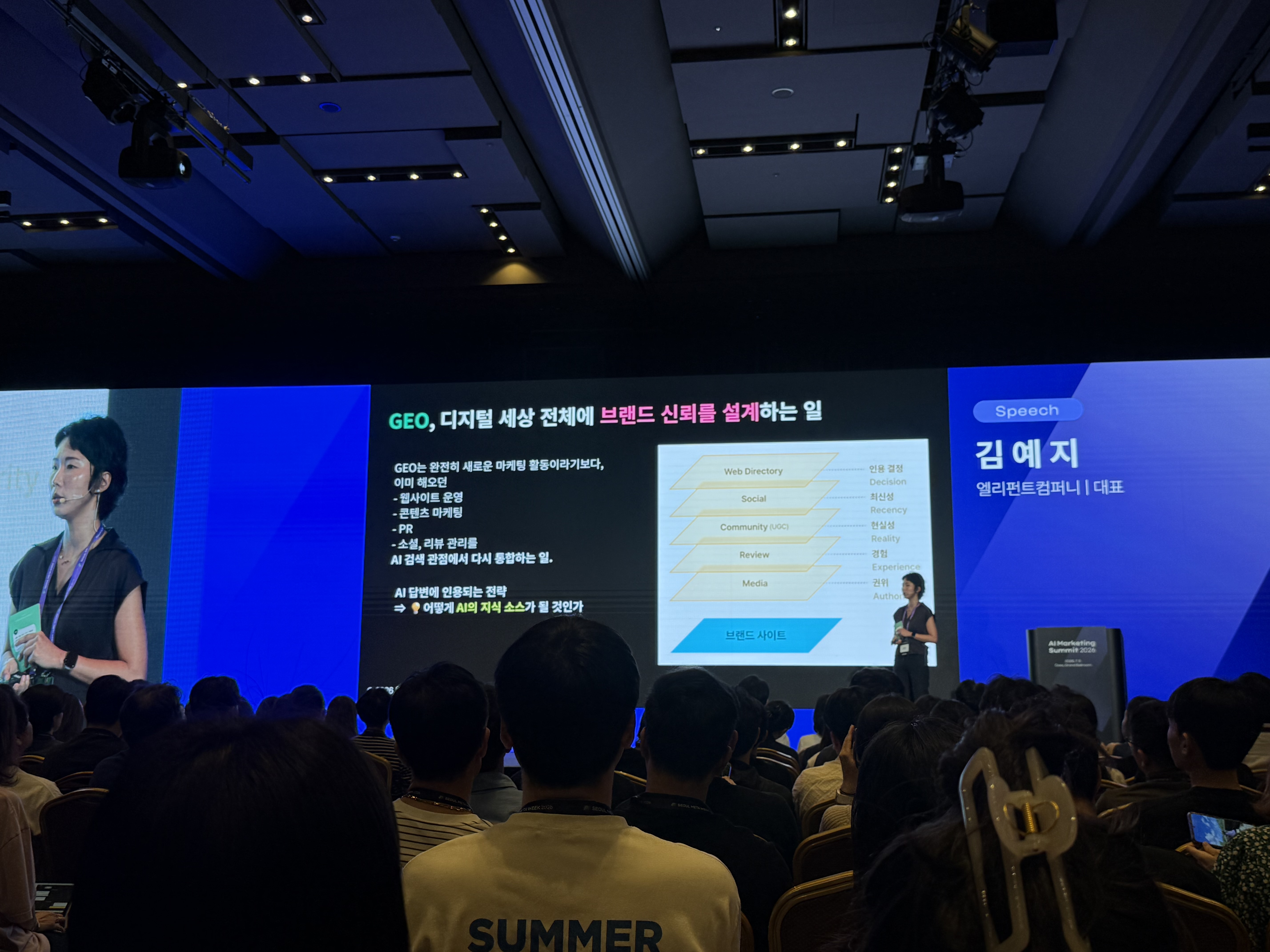 서울메타위크 AI Marketing Summit 2026 GEO 세션 현장. 발표 화면에는 “GEO, 디지털 세상 전체에 브랜드 신뢰를 설계하는 일”이라는 문구와 함께 브랜드 사이트, 미디어, 리뷰, 커뮤니티, 소셜, 웹 디렉토리 등이 신뢰 신호로 쌓이는 구조가 설명되어 있다.