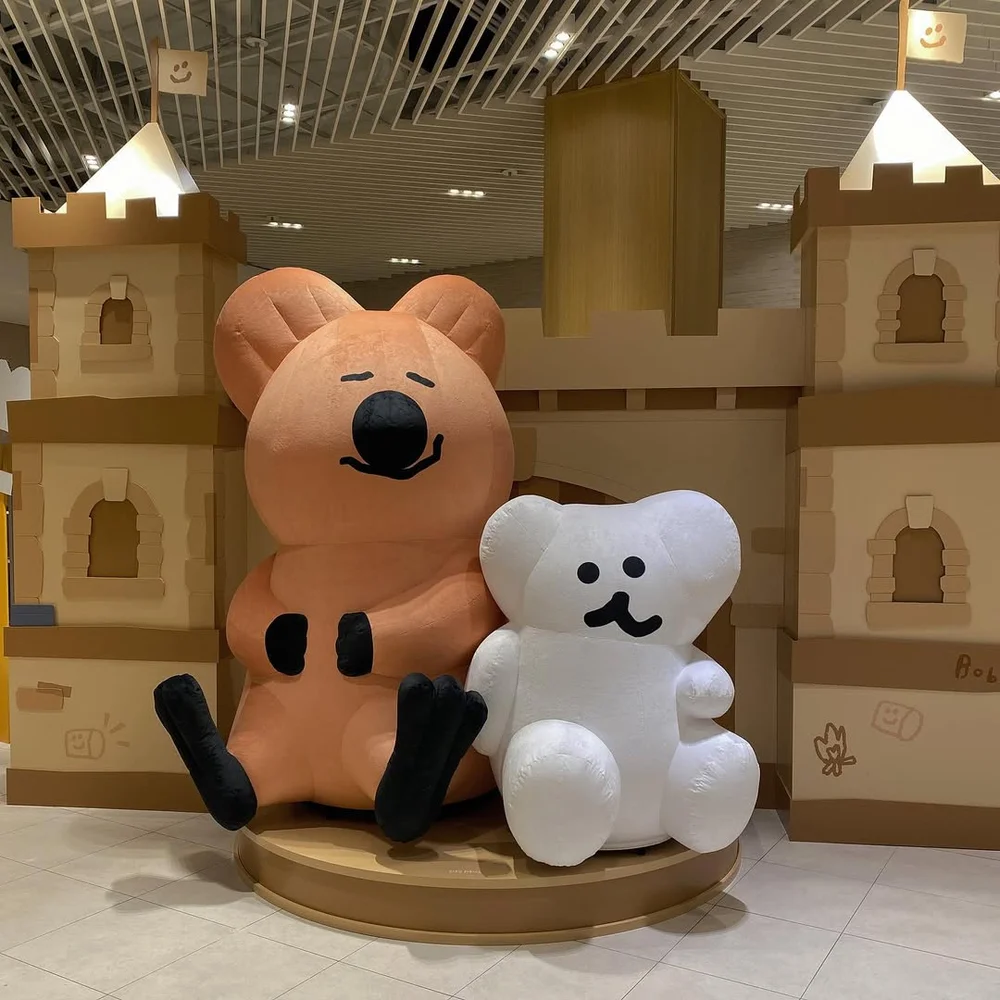 골판지 상자로 만든 성 앞에 커다란 갈색 쿼카 조형물과 하얀 보보 조형물이 나란히 앉아 있는 전시장 모습